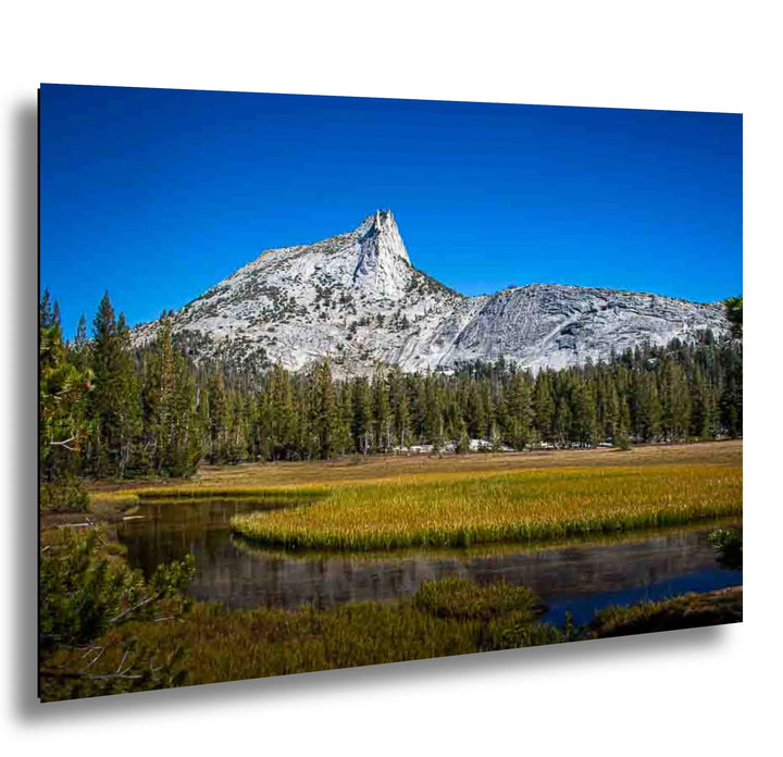 Scenic view of a towering mountain peak with lush green forest and a calm stream flowing through golden fields under a bright blue sky.