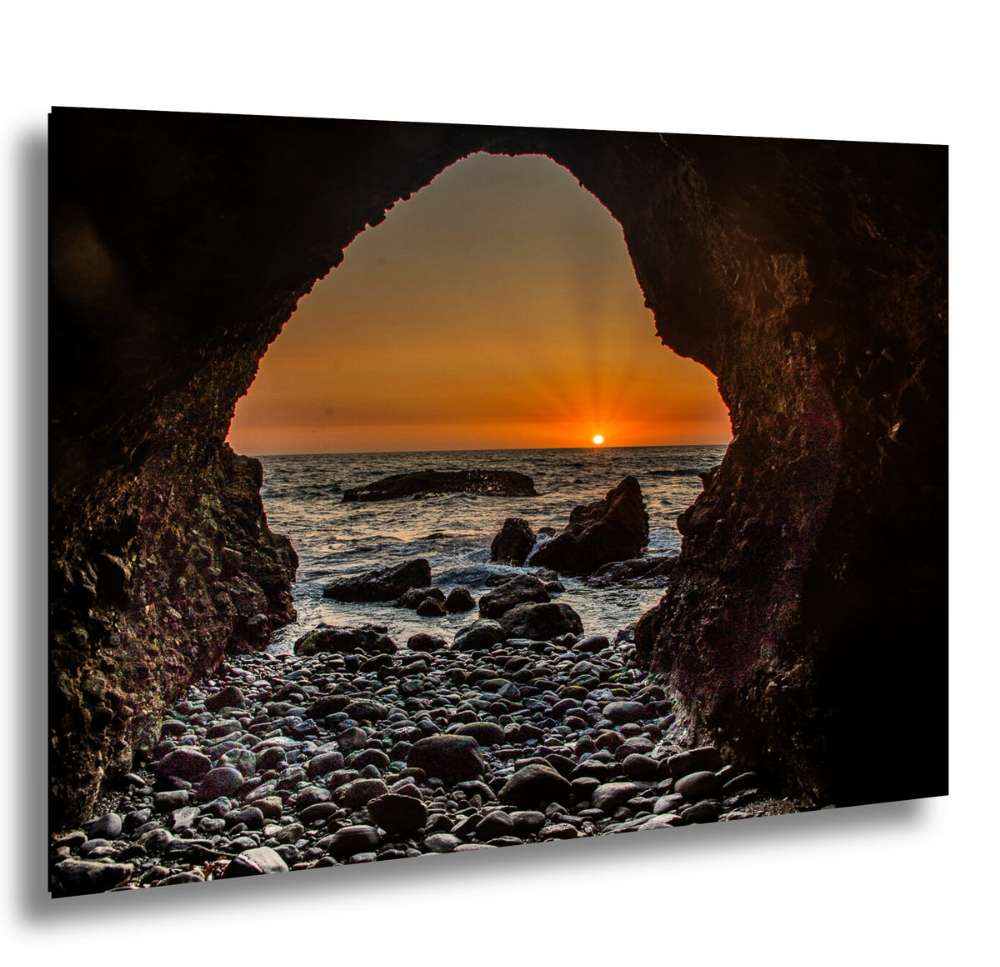 Sunset view through a rocky cave opening, with rays of light shining over the ocean and pebbled beach below.