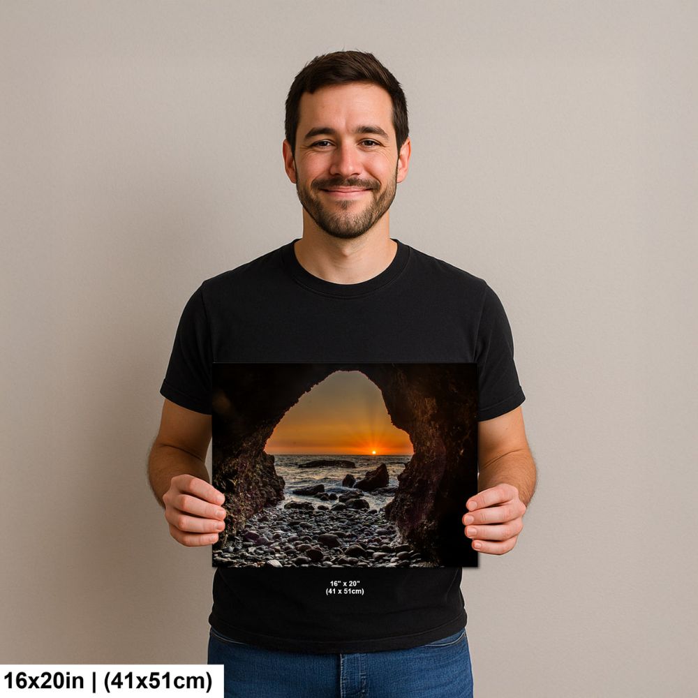 Man holding a 16x20 inch print of a sunset through a rocky cave opening, with the ocean and pebbled beach visible in the distance.